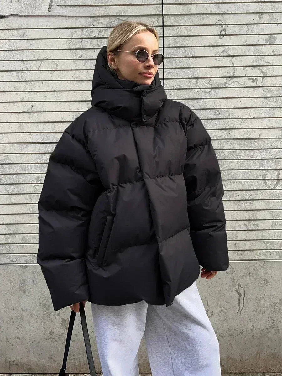 Vrouw draagt een stijlvolle gewatteerde winterjas, ideaal voor koude dagen. De jas is oversized en comfortabel, met een royale capuchon voor extra warmte en bescherming tegen de kou. Ze combineert de jas met een gebreide muts, zonnebril en casual outfit, wat een modieuze en warme uitstraling creëert. Perfect voor winterse wandelingen en dagelijkse activiteiten in de kou.