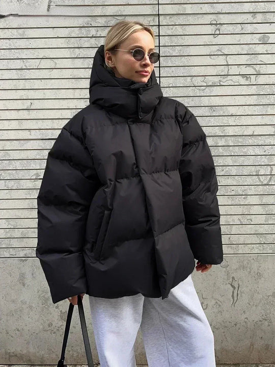 Vrouw draagt een stijlvolle gewatteerde winterjas, ideaal voor koude dagen. De jas is oversized en comfortabel, met een royale capuchon voor extra warmte en bescherming tegen de kou. Ze combineert de jas met een gebreide muts, zonnebril en casual outfit, wat een modieuze en warme uitstraling creëert. Perfect voor winterse wandelingen en dagelijkse activiteiten in de kou.