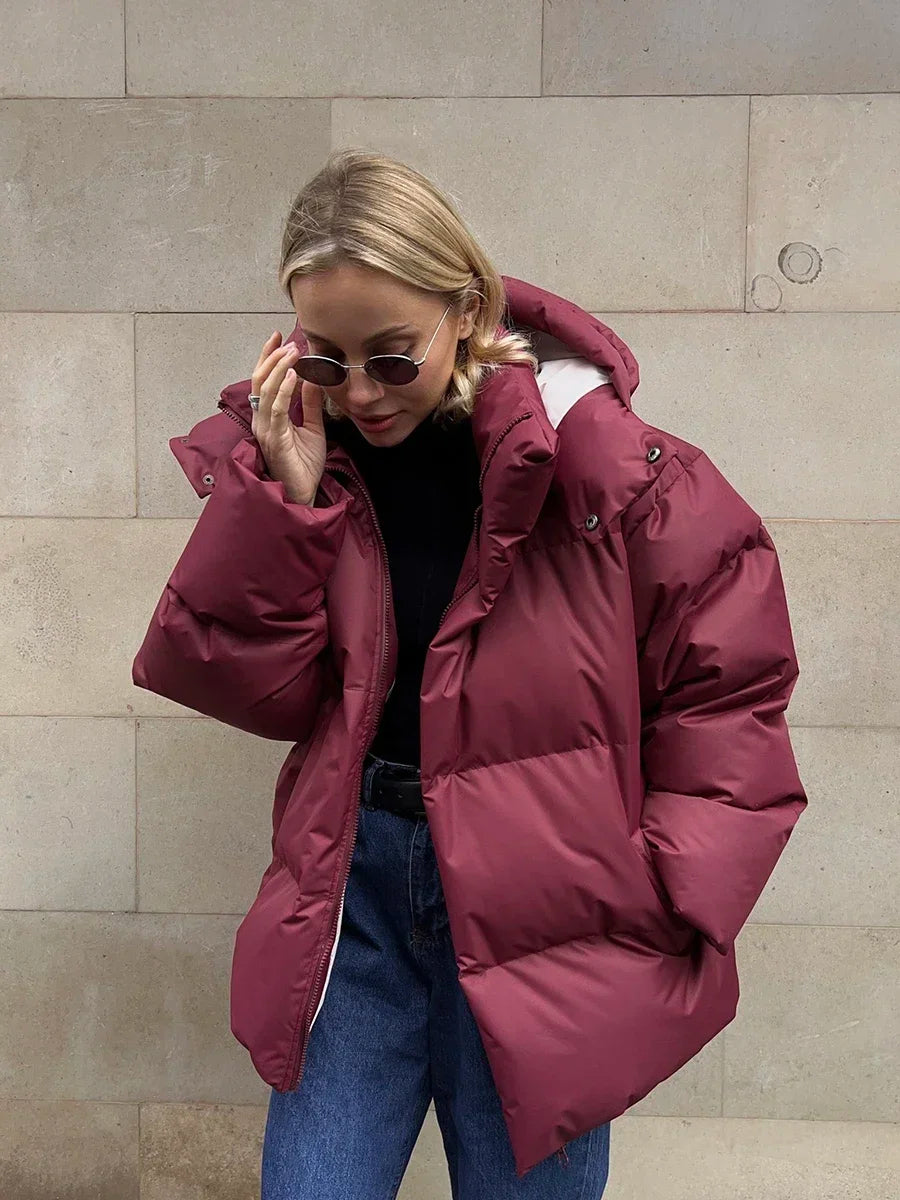 Vrouw draagt een stijlvolle gewatteerde winterjas, ideaal voor koude dagen. De jas is oversized en comfortabel, met een royale capuchon voor extra warmte en bescherming tegen de kou. Ze combineert de jas met een gebreide muts, zonnebril en casual outfit, wat een modieuze en warme uitstraling creëert. Perfect voor winterse wandelingen en dagelijkse activiteiten in de kou.