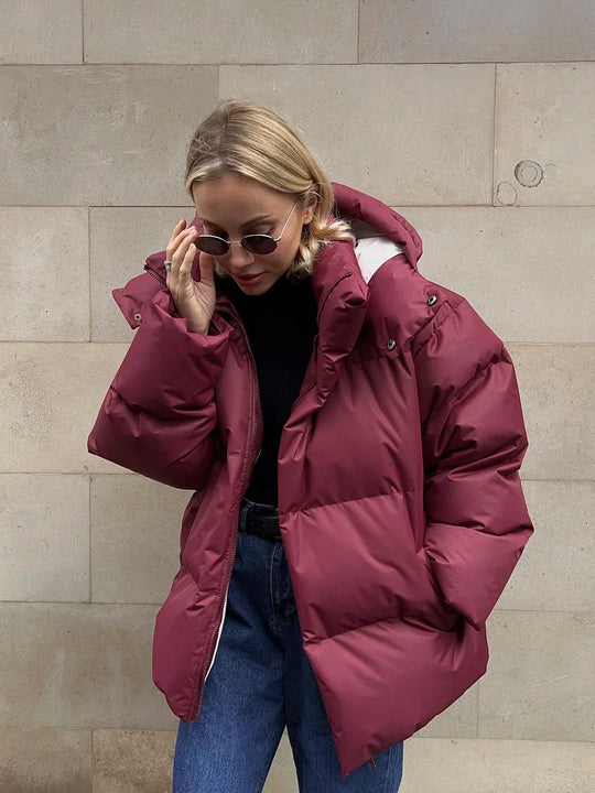 Vrouw draagt een stijlvolle gewatteerde winterjas, ideaal voor koude dagen. De jas is oversized en comfortabel, met een royale capuchon voor extra warmte en bescherming tegen de kou. Ze combineert de jas met een gebreide muts, zonnebril en casual outfit, wat een modieuze en warme uitstraling creëert. Perfect voor winterse wandelingen en dagelijkse activiteiten in de kou.