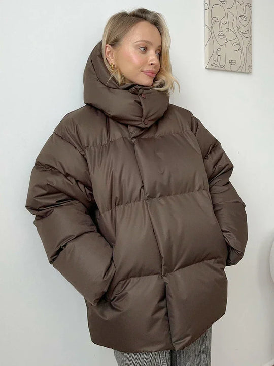 Vrouw draagt een stijlvolle gewatteerde winterjas, ideaal voor koude dagen. De jas is oversized en comfortabel, met een royale capuchon voor extra warmte en bescherming tegen de kou. Ze combineert de jas met een gebreide muts, zonnebril en casual outfit, wat een modieuze en warme uitstraling creëert. Perfect voor winterse wandelingen en dagelijkse activiteiten in de kou.