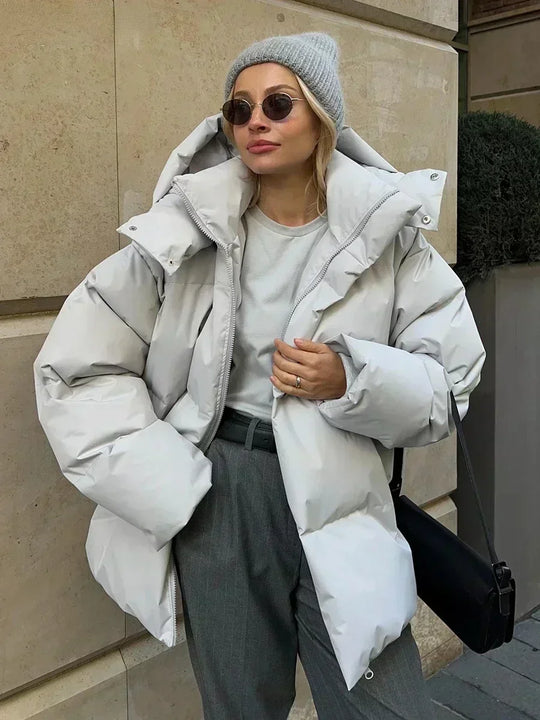 Vrouw draagt een stijlvolle gewatteerde winterjas, ideaal voor koude dagen. De jas is oversized en comfortabel, met een royale capuchon voor extra warmte en bescherming tegen de kou. Ze combineert de jas met een gebreide muts, zonnebril en casual outfit, wat een modieuze en warme uitstraling creëert. Perfect voor winterse wandelingen en dagelijkse activiteiten in de kou.