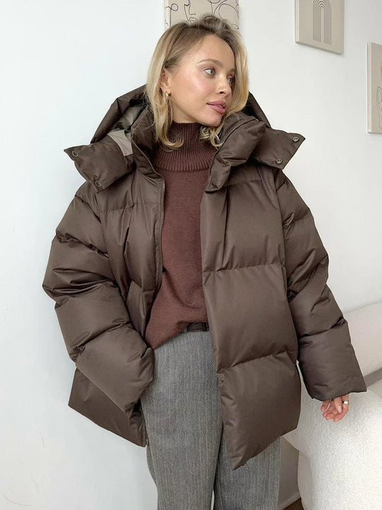 Vrouw draagt een stijlvolle gewatteerde winterjas, ideaal voor koude dagen. De jas is oversized en comfortabel, met een royale capuchon voor extra warmte en bescherming tegen de kou. Ze combineert de jas met een gebreide muts, zonnebril en casual outfit, wat een modieuze en warme uitstraling creëert. Perfect voor winterse wandelingen en dagelijkse activiteiten in de kou.